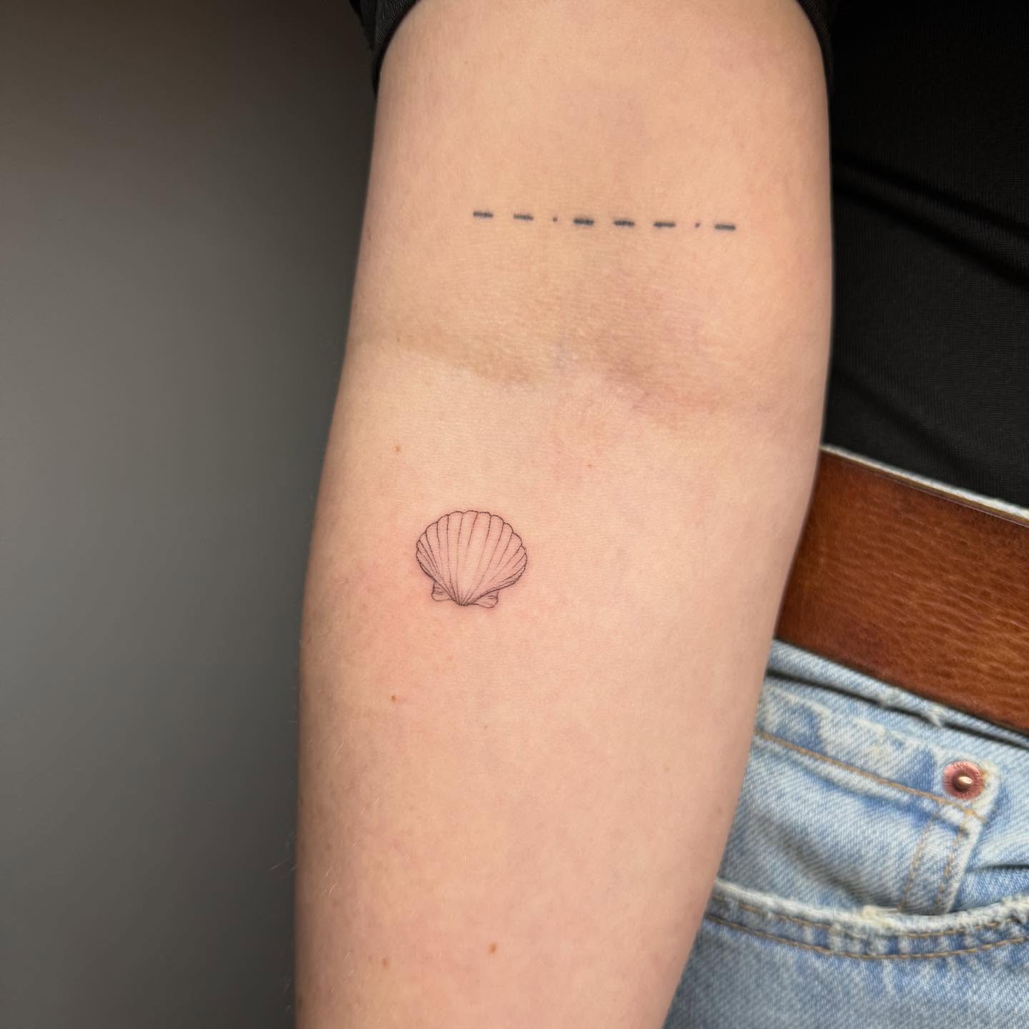 Small fine-line seashell tattoo on an upper arm by Luna Fino